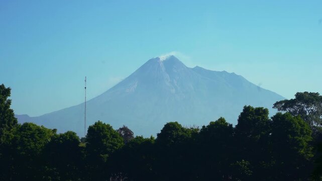 Mount Merapi rises beyond a dark green treeline near Prambanan Temple under a hazy tropical sky.