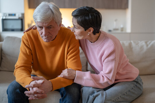 Senior couple sitting on sofa at home, emphatic elderly woman comforts upset overwhelmed husband, worried wife provides support and embracing thoughtful stressed partner, family unity and love.