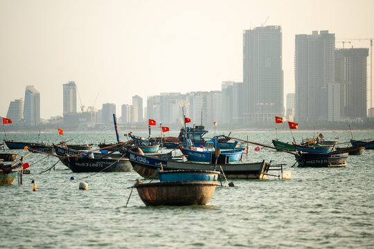 Various Vietnamese boats for seafood fishing and transportation at bay with equipment on board, traditional local fishermen fleet with round basket boats on water against modern city skyscrapers.