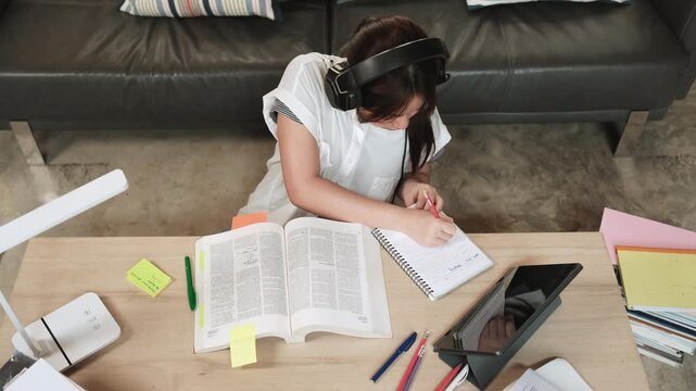 Asian teenage girl in headphones concentrates on learning at home, reading textbooks, note writing, self-tutorial exams, preparation for back to school, Homeschooling, university education lifestyle.