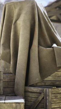 In a dimly lit corner of a warehouse, a covered statue stands shrouded in a large cloth. Surrounding wooden crates add to the atmosphere of secrets and forgotten stories.