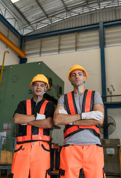 Two male workers in orange vests and hard hats stand arms crossed before a green power press machine beneath roof trusses