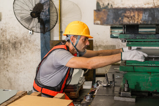 Male machine operator in an orange vest leans forward, reaching gloved hands into a green power press to adjust its die