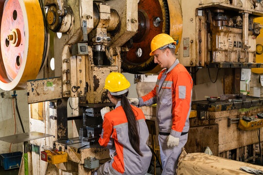 Male worker guiding colleague's hand while operating metal stamping die press together supporting efficient production and workplace safety inside a busy metalworking factory setting
