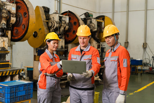 Three workers in yellow hard hats and orange coveralls gather around a laptop beside power press machines on the factory floor