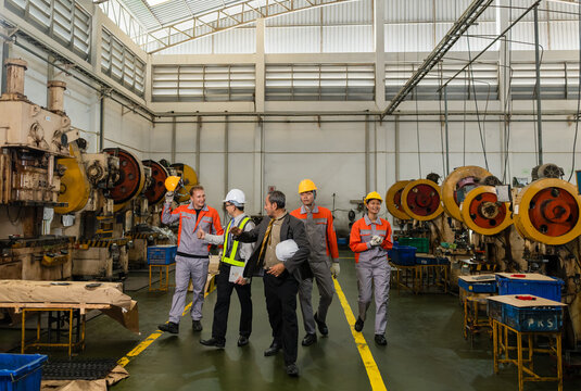 A manager, an engineer holding a tablet, and factory workers walk down an aisle between rows of metal stamping presses
