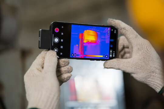 Close up of gloved hands holding thermal imaging phone attachment showing motor heat map supporting efficient production and workplace safety inside a busy metalworking factory setting