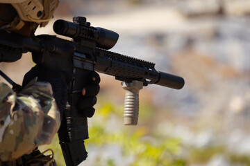 Obraz premium Close-up of a soldier in camouflage gear holding an assault rifle with a scope and vertical foregrip during a simulated tactical military combat training session on an outdoor sunny battlefield.