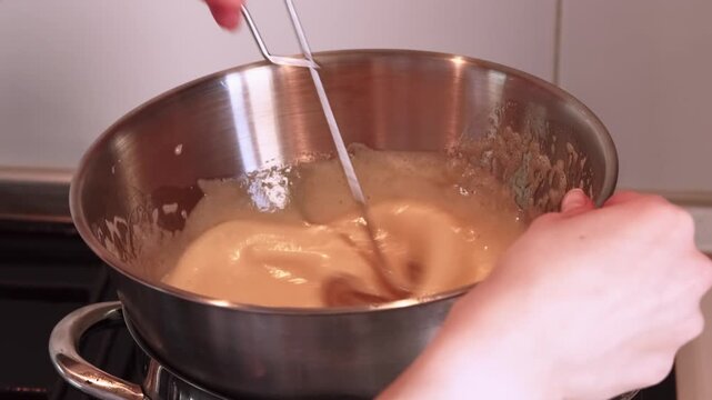 Cooking meal. Whisking process. Chef hand stirring orange foam cream liquid in stainless steel bowl with spring tool on heating stove.