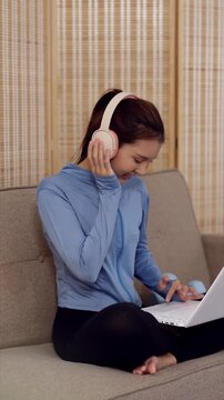 Young Asian woman wearing pink headphones working on laptop while sitting in yoga outfit on living room couch