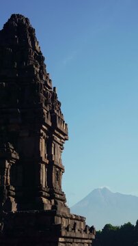 Shadowed Prambanan Temple tower forms a dramatic silhouette beside distant Mount Merapi under clear tropical skies.