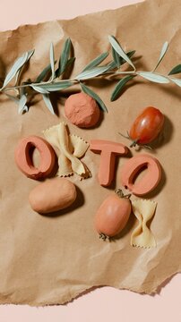 Terracotta clay letters spelling the word orto on wrinkled brown paper with fresh tomatoes, farfalle pasta, and an olive branch for food branding