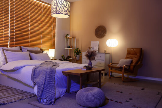 Comfortable bed, wooden bench and glowing lamps in bedroom at night