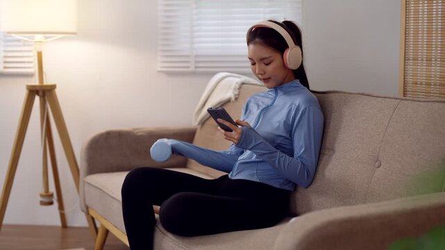 Young Asian woman in sportswear listening to music through wireless headphones while using smartphone on living room sofa