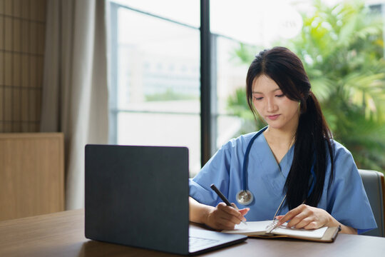 Female asian healthcare worker wearing blue scrubs sitting while confidently using a laptop and writing note near window in a modern clinic or hospital setting, representing innovation in healthcare