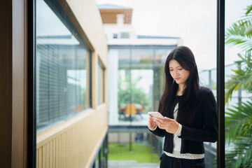 Fototapeta premium A focused young Asian woman in casual business attire checks her smartphone on a contemporary balcony, ideal for technology and lifestyle campaigns