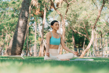 Fototapeta premium Asian woman meditating in a peaceful park setting, sitting cross-legged on green grass with trees in the background on a sunny day. Concept of mindfulness, self-care, and outdoor relaxation