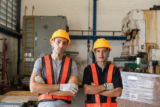 Two male workers in orange vests and hard hats stand arms crossed inside a metalworking machine shop near rusted power press