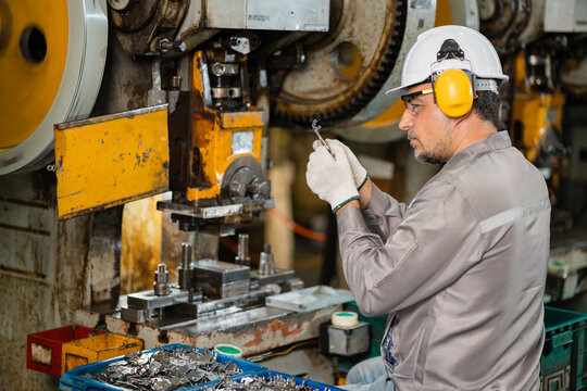 Technician inspecting small stamped metal bracket beside punch press die close up supporting efficient production and workplace safety inside a busy metalworking factory setting