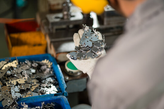 Worker's gloved hand holding cluster of stamped metal brackets above blue parts bin
