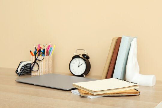 Modern school desk with laptop, alarm clock and stationery in room near beige wall