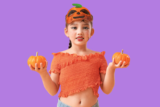 Young girl in pumpkin mask and orange top holding two small pumpkins for halloween celebration, She has playful face paint and a braided hairstyle