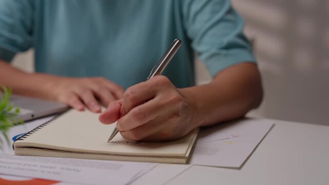 Asian woman works from home, surrounded by notes, a laptop, and messages. She browses the internet, writes ideas, and researches for her startup, utilizing wireless technology.
