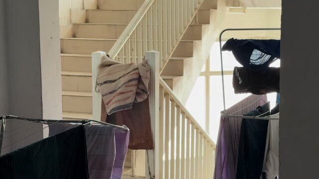 A static shot looking through a doorway at wet clothes hanging on drying racks inside an apartment building stairwell. Bright daylight streams through the background window illuminating the stairs.