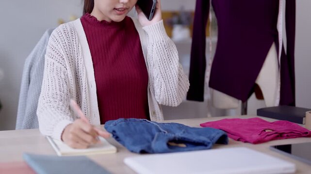 Young Asian entrepreneur taking phone order while writing notes surrounded by clothing in workspace