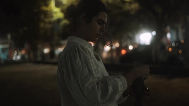Woman standing outdoors at night adjusting item in hands under streetlights