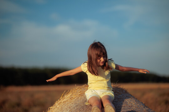 Joyful Little Girl Standing On Straw Bale Holding Arms Like Flying. Happy young child pretends to fly while playing outdoors on wheat hay