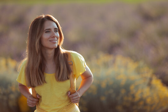 Smiling Woman In Yellow Dress And Backpack Looking Aside In Flower Field. Cheerful lady wears a matching outfit posing happily inside vibrant yellow meadow