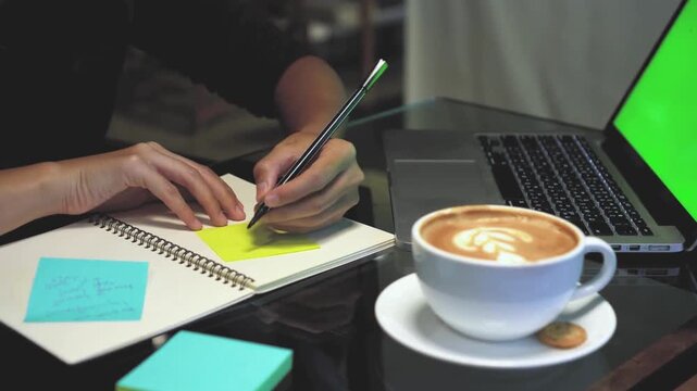 Woman hands sticking yellow note on spiral notebook while working on laptop with green screen chroma key in cozy cafe setting with hot cup of latte coffee.