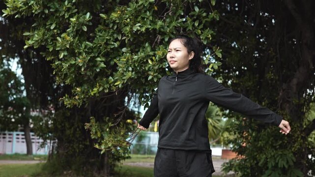 Asian young woman in black sportswear doing cross-body shoulder arm stretch warm-up exercise under green trees in park before outdoor running workout for health.