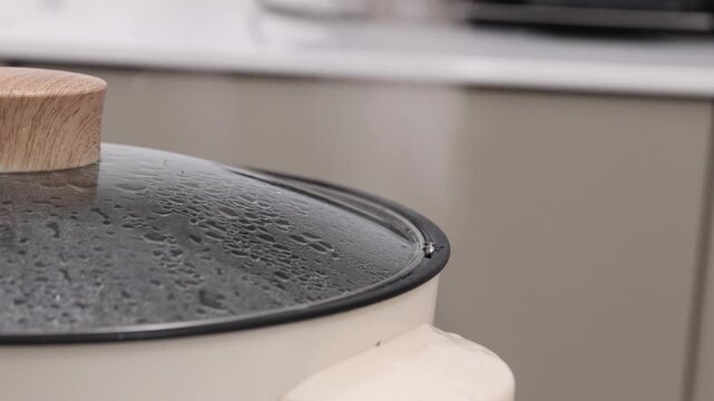 Close-up shot of an electric skillet or hot pot lid with boiling water inside, heavy condensation droplets on the glass, and steam escaping from the rim.