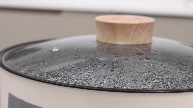 Close-up shot of an electric skillet or hot pot lid with boiling water inside, heavy condensation droplets on the glass, and steam escaping from the rim.