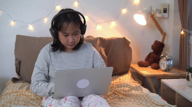 Focused Young Girl sits comfortably on bed using a laptop wearing headphones Online in Cozy Bedroom