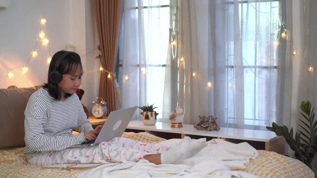 Young Girl wearing wireless headphones Studying on Laptop from Bed in a Cozy Evening Bedroom