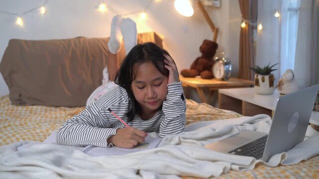 Young girl is  lies in bed, writing homework diligently in cozy bedroom 