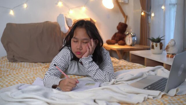 Exhausted young girl fell asleep while doing her homework in bed with notebook and laptop in warmly lit bedroom