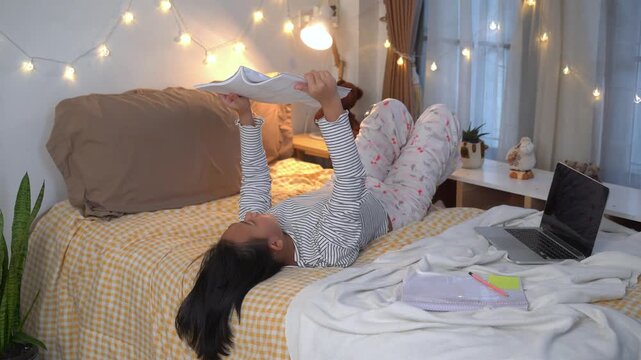 Young woman lying on bed reading a textbook, study materials and a laptop, Comfortable Home Study Session on a Cozy Evening
