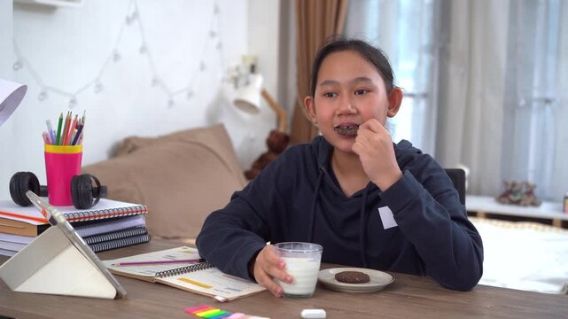 Young girl happily enjoys a break with milk and cookies during online lessons at home