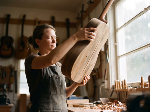 Female luthier inspecting handmade wooden acoustic guitar finish by window light, craftsman woodworking art and music instrument manufacturing lifestyle concept