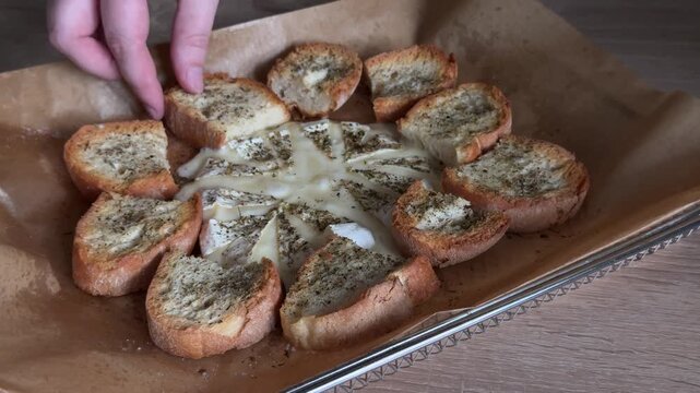 Baked brie cheese with toasted bread and melted cheese stretching from a crispy slice, close-up of warm appetizer with herbs, rustic homemade food, savory snack and entertaining concept