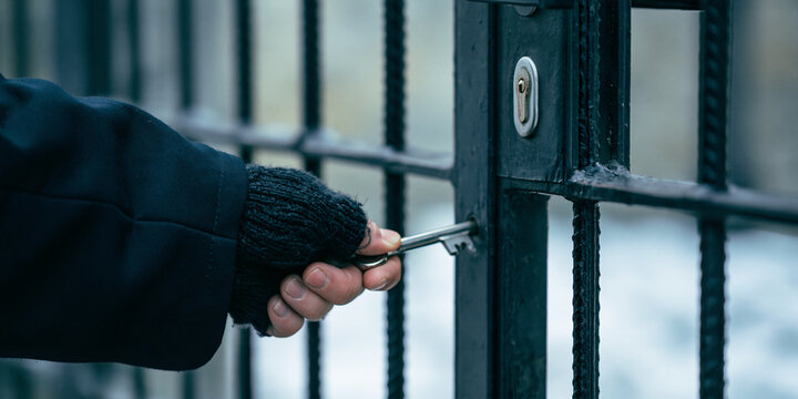 A person's hand wearing a glove is inserting a key into a metal gate lock. The gate appears to be made of black metal bars, and the keyhole is visible.