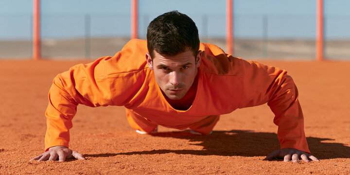 A man in an orange jumpsuit performs a push-up outdoors in an orange-toned environment. He is looking directly at the camera with a determined expression. The ground appears to be sand or dirt