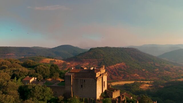 High-angle drone shot capturing a medieval stone castle in the foreground, with firefighting Canadair aircraft flying in the distance over mountain ridges toward a rising forest fire smoke plume.
