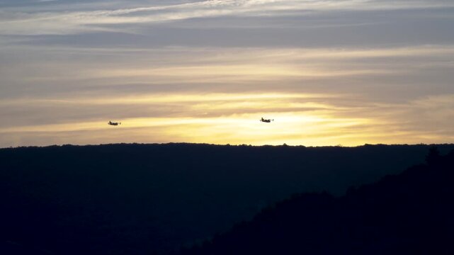 Beautiful cinematic wide shot of three firefighting Canadair aircraft flying in formation, silhouetted against a dramatic golden sunset sky over the dark mountain ridges of the Salagou Lake area