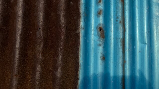 Close-up of weathered corrugated metal, half rusty brown and half painted blue. Dramatic texture, erosion, and corrosion, showing old age and decay