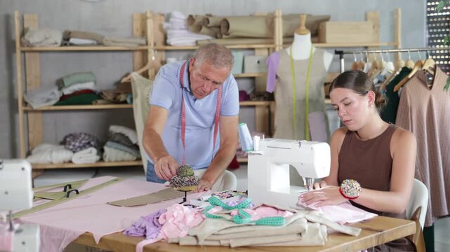 Working day in sewing workshop, textile industry. Girl master sew clothes in sewing machine, mature man pins paper pattern to fabric, attaches template to fabric, preparing to cut fabric into pieces.